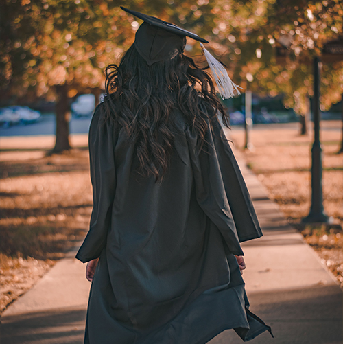 A photo of a girl with her back to the camera, in a graduation gown walking in a park.
